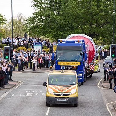 Large truck transporting a Rolls Royce Aero Engine, with a crowd watching from the roadside. Convoi Exceptionnel Escort vehicle.